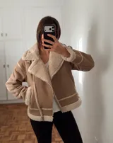 Campera tipo gamulán de gamuza sintética color tostado, con interior y solapas de corderito sintético color crema. Presenta cierre frontal cruzado, bolsillos laterales y detalles de costuras reforzadas.