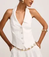 Chaleco sastre de frente única, color blanco, con cuello solapa, cierre frontal con botones y ruedo en punta en forma de V. Presenta un diseño entallado y espalda descubierta.