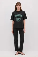 Remera negra de algodón de manga corta y cuello redondo, con diseño holgado y estampado frontal de estilo universitario que dice 'NEW YORK CITY' y 'BROOKLYN' en color verde.