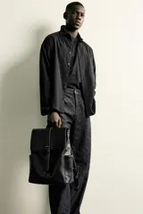 Mochila de diseño minimalista en color negro, confeccionada en material sintético con acabado liso. Presenta una solapa frontal con cierre de hebilla decorativa y sistema de imán, compartimento principal con cremallera, bolsillos laterales externos, asa superior de mano y correas de hombro ajustables y acolchadas.