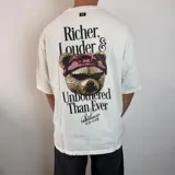 Remera de corte oversize color blanco, con estampa gráfica en la espalda que presenta un oso con anteojos y bandana, acompañada de la frase 'Richer, Louder & Unbothered Than Ever'.