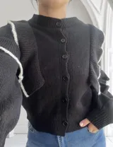Cárdigan negro de punto con cuello redondo, cierre frontal con botones y volados blancos en los hombros.