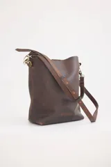 Cartera tipo bucket bag de cuero marrón con acabado rústico, correa larga ajustable y herrajes metálicos en tono bronce.