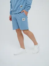 Bermuda de felpa de algodón color celeste, con bolsillos laterales y detalle de costuras verticales al frente. Presenta un parche distintivo de la marca en la pierna izquierda.