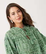 Blusa tipo bata de algodón con tejido laise bordado, cuello redondo con cordones y borlas, y mangas largas abullonadas con puños ajustados.