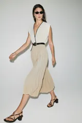 Vestido midi bicolor con parte superior blanca de escote en V cruzado con ribete negro y falda plisada en color tostado. Cuenta con mangas sisa, cintura elástica y un cinturón negro tipo reptil con hebilla ovalada.