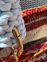 Bolso tejido a mano con hilo de trapillo, eco-cuero y textiles, con un diseño central de querubín coronado y alas, sobre un fondo de franjas rojas, blancas y azules. Posee asas cortas tejidas en color celeste claro.