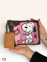 Sobre de cuero marrón con diseño pintado a mano que presenta al personaje Snoopy sobre un fondo rosado y la palabra 'heal'. Cuenta con cierre superior y etiqueta de cuero colgante.