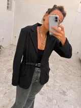 Blazer negro de largo medio con solapas clásicas, sin cruce frontal y con cinto de cuero fino incorporado en la cintura para ajuste.