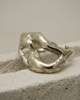 Brazalete abierto de diseño orgánico y voluminoso, con acabado metálico brillante. Su forma irregular y ergonómica permite un ajuste cómodo en la muñeca o antebrazo.