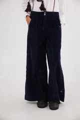 Pantalón sastrero de tiro medio confeccionado en pana de algodón. Presenta un diseño de pierna ancha con costuras verticales decorativas, bolsillos delanteros y traseros, y un detalle de aplique con ojalillo metálico en el ruedo de una de las piernas.