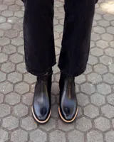 Botinetas tipo Chelsea de cuero negro, con paneles laterales elásticos y detalle de textura grabada en el talón. Presentan una suela plana con plataforma de madera clara y base de goma negra.