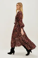 Vestido midi de corte holgado con estampado ikat en tonos bordó y tierra. Presenta cuello redondo con lazos, mangas largas abullonadas con puños elásticos y falda con volados escalonados. Incluye un cinturón ancho de estilo rústico con tachas y flecos.