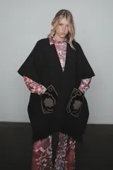 Ruana de tejido de punto color negro, con diseño abierto, flecos en el ruedo y bolsillos frontales destacados con bordados artesanales en tonos contrastantes.