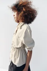 Chaqueta estilo safari color beige, con cuello de solapa tejido en crochet, manga larga, bolsillos delanteros con solapa y cintura ajustable con cordones. Cierre frontal con botones metálicos a presión.