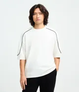 Remera de algodón color blanco, con cuello redondo, manga corta y detalle de raya negra contrastante que recorre la parte superior de los hombros y mangas. Diseño de corte holgado.
