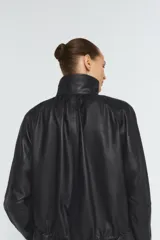 Chaqueta negra de piel 100% con cuello alto, manga larga con puño, bolsillos delanteros y cintura ajustable con cordón elástico y stopper. Cierre frontal con cremallera metálica y botones a presión ocultos por solapa.