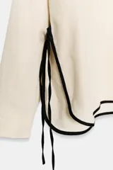 Camisa de lino color crudo con cuello solapa, manga larga y bajo asimétrico. Presenta detalles de ribetes negros a contraste y aberturas laterales con tiras para ajustar.