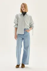 Camisa a rayas verticales de calce recto, con cuello clásico y mangas abullonadas con detalles de frunces y lazos en los puños. Presenta un pequeño bordado en el pecho.