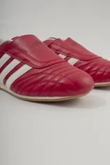 Championes Adidas rojos de cuero sintético, diseñados para taekwondo. Presentan el logo de Adidas en la lengüeta y las tres rayas blancas características en los laterales. El interior es acolchado y de color blanco.