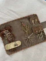 Set de accesorios compuesto por un sobre organizador de cuero gamuzado marrón con costuras decorativas, que incluye un conjunto de piezas de joyería artesanal en metal dorado: un broche, un collar de diseño geométrico con cordones de cuero, y tres pares de caravanas con diseños abstractos y orgánicos.
