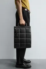 Bolso tipo shopper de diseño acolchado con patrón de cuadros, confeccionado en material sintético negro. Presenta asas de mano integradas en la estructura superior y un detalle de borla colgante con herraje metálico.
