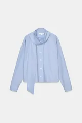 Camisa de algodón color celeste, con cuello alto y lazo integrado en el mismo tejido. Presenta cierre frontal con botones, manga larga con puños y un bolsillo frontal en el pecho. Diseño de corte recto.