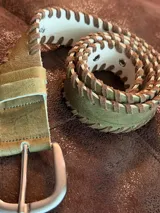 Cinturón de cuero color verde pistacho con detalles entrelazados en cuero gamuza color cocoa. Presenta hebilla metálica plateada y ojales reforzados.
