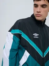 Campera deportiva Umbro modelo Beat, confeccionada en nylon ligero con diseño de bloques de color en blanco, azul marino y verde azulado. Presenta cierre frontal, cuello alto y elásticos en puños y cintura.