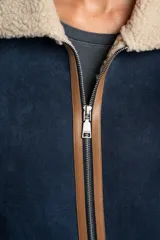 Campera tipo gamulán de corte oversize, color azul marino con interior y cuello de corderito en tono beige. Presenta detalles de costuras y bordes en cuero sintético color marrón, cierre frontal y mangas largas.