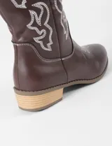 Botas texanas de caña media en color marrón oscuro, confeccionadas en cuero vegano. Presentan bordados decorativos en hilo claro en la caña y en la puntera, con un taco bajo de madera y suela de caucho.