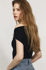 Top de punto fino color negro, con diseño de un solo hombro y escote asimétrico. Presenta un panel fruncido en el hombro con un adorno metálico dorado y drapeado lateral en la cintura que aporta un efecto entallado.