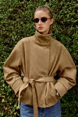 Chaqueta de gamuza sintética color marrón, con cuello subido, cierre frontal cruzado con botones y mangas largas anchas. Cuenta con bolsillos delanteros y cinturón ajustable del mismo tejido.