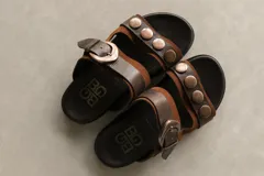 Sandalias bajas de cuero marrón con doble tira, hebilla ajustable y apliques de tachas metálicas.