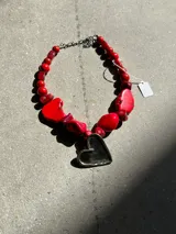Collar artesanal con cadena de alpaca, compuesto por cuentas de coral rojo de forma irregular y cuentas redondas más pequeñas. Incluye un dije grande en forma de corazón de metal plateado.