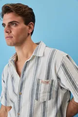 Camisa de manga corta a rayas verticales en tonos azul y blanco, con cuello camisero, cierre frontal de botones y un bolsillo en el pecho con etiqueta aplicada.