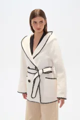 Chaqueta blanca de corte holgado con capucha, cierre frontal cruzado con botones y cinturón de tela ajustable. Presenta detalles de costuras decorativas en color negro en los bordes, solapas, bolsillos y cinturón.