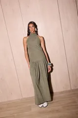 Vestido largo holgado color verde oliva, con escote halter y cuello alto con volado. Presenta un corte con fruncido a la altura de la cadera que genera un maxi volado en la falda.