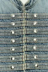 Chaqueta denim entallada de cuello redondo y manga larga, en tono azul desgastado. Presenta un diseño inspirado en estilos militares con múltiples filas de botones metálicos plateados en el frente y cierre con ganchos ocultos.