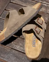 Sandalias tipo zueco marca Rivaro, confeccionadas en cuero gamuzado color beige. Presentan dos tiras anchas con hebilla metálica, plantilla anatómica de corcho y látex, y suela de goma antideslizante.