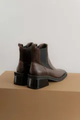 Botas tipo chelsea de cuero color marrón oscuro con acabado brillante. Presentan punta cuadrada con detalle de puntera metálica, elásticos laterales y taco cuadrado bajo.