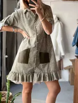 Vestido camisero de corte recto y largo por encima de la rodilla, confeccionado en pana elastizada con estampado floral pequeño. Presenta cuello camisero, cierre frontal con botones, mangas tres cuartos con puños vueltos, bolsillos frontales de cuero en contraste y ruedo con volado.