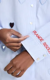 Camisa de algodón a rayas celestes y blancas, con cuello clásico, cierre frontal de botones, mangas largas y aberturas troqueladas en forma de corazón junto a bordados decorativos y texto en el hombro.
