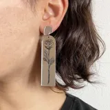 Aros colgantes largos de acrílico transparente con diseño de rosa grabada en blanco y pieza superior cuadrada de color blanco.