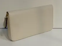 Billetera rectangular de cuero color beige con cierre perimetral y tirador de cuero. Cuenta con múltiples compartimentos internos para tarjetas, documentos y billetes, además de un bolsillo interno con cierre para monedas y forro textil en color rojo.