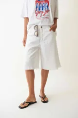 Bermuda de denim blanco de tiro bajo y corte holgado, con detalles desgastados y terminación desflecada en las perneras. Cuenta con trabillas en la cintura y cierre frontal con botón y cremallera.