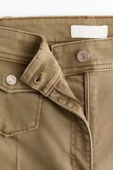 Pantalón de sarga color tostado, de tiro alto y corte acampanado con diseño tobillero. Presenta bolsillos frontales tipo parche con solapa abotonada y pliegues marcados al frente y en la espalda.