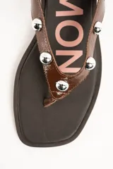 Sandalias tipo ojota de cuero marrón con acabado charol, con tiras decoradas con tachas metálicas esféricas y suela plana.