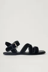 Sandalia plana de cuero negro, con diseño minimalista de tiras finas cruzadas sobre el empeine y una tira ajustable con hebilla metálica en el tobillo.