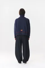 Buzo de tejido de punto en color azul marino, con cuello alto y cierre frontal parcial. Presenta un diseño con franjas en color marrón y detalles de líneas en rojo en el pecho, hombros y ruedo. Tiene corte holgado y mangas largas con puños acanalados.